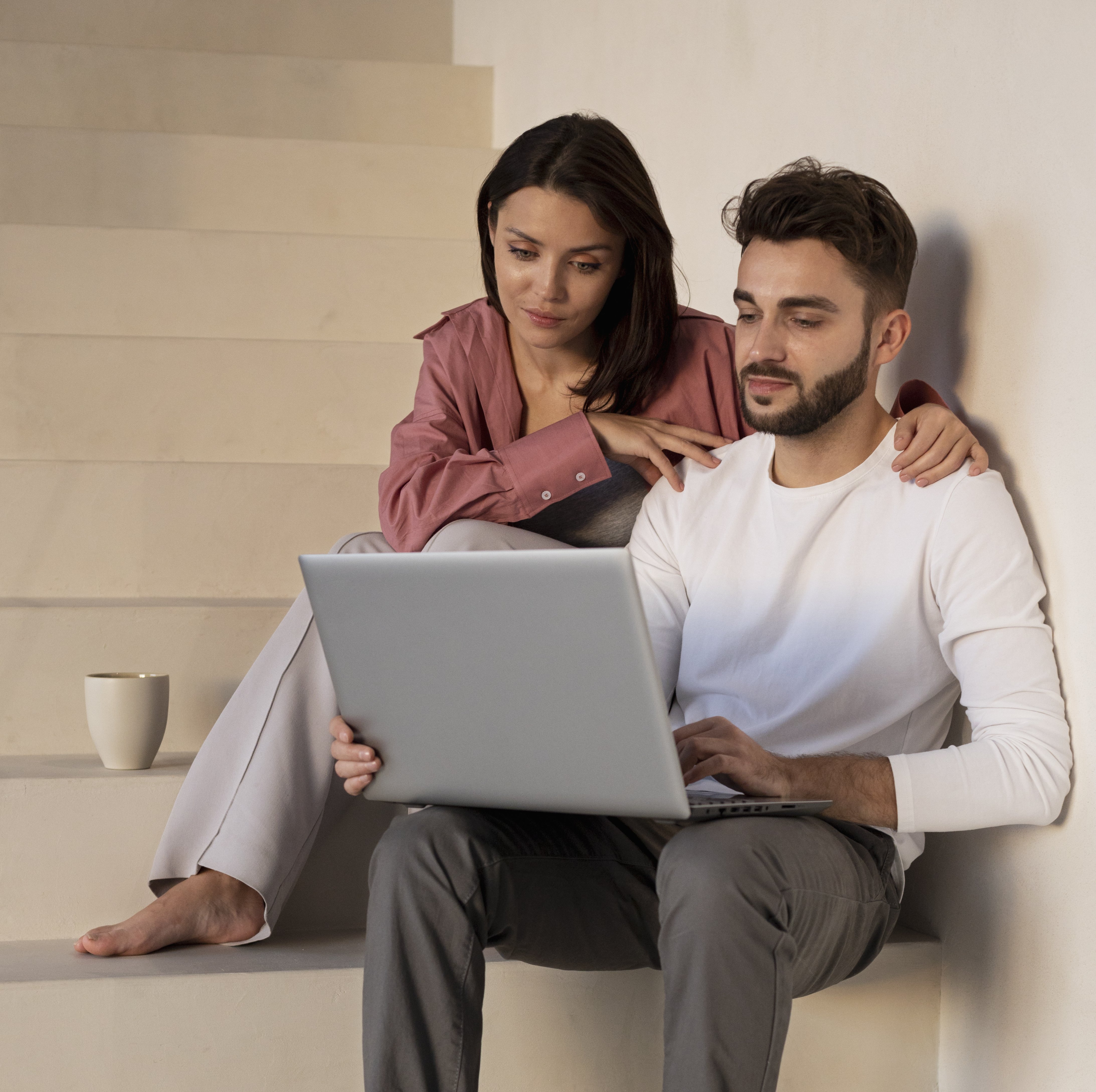 Couple booking online venue, sitting on staircase, looking at laptop screen. Male smiling, female enjoying coffee. Website's about image. 